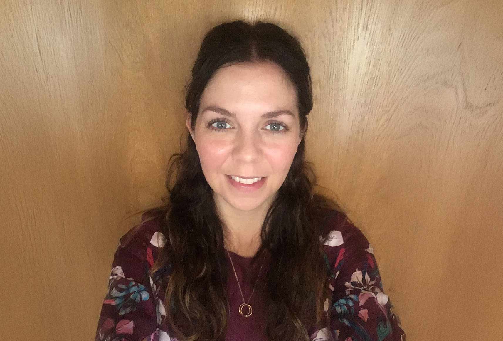 A person with long, wavy dark hair is smiling at the camera. They are wearing a patterned top with floral designs and a necklace with two interlocking rings. The background is a wooden surface.