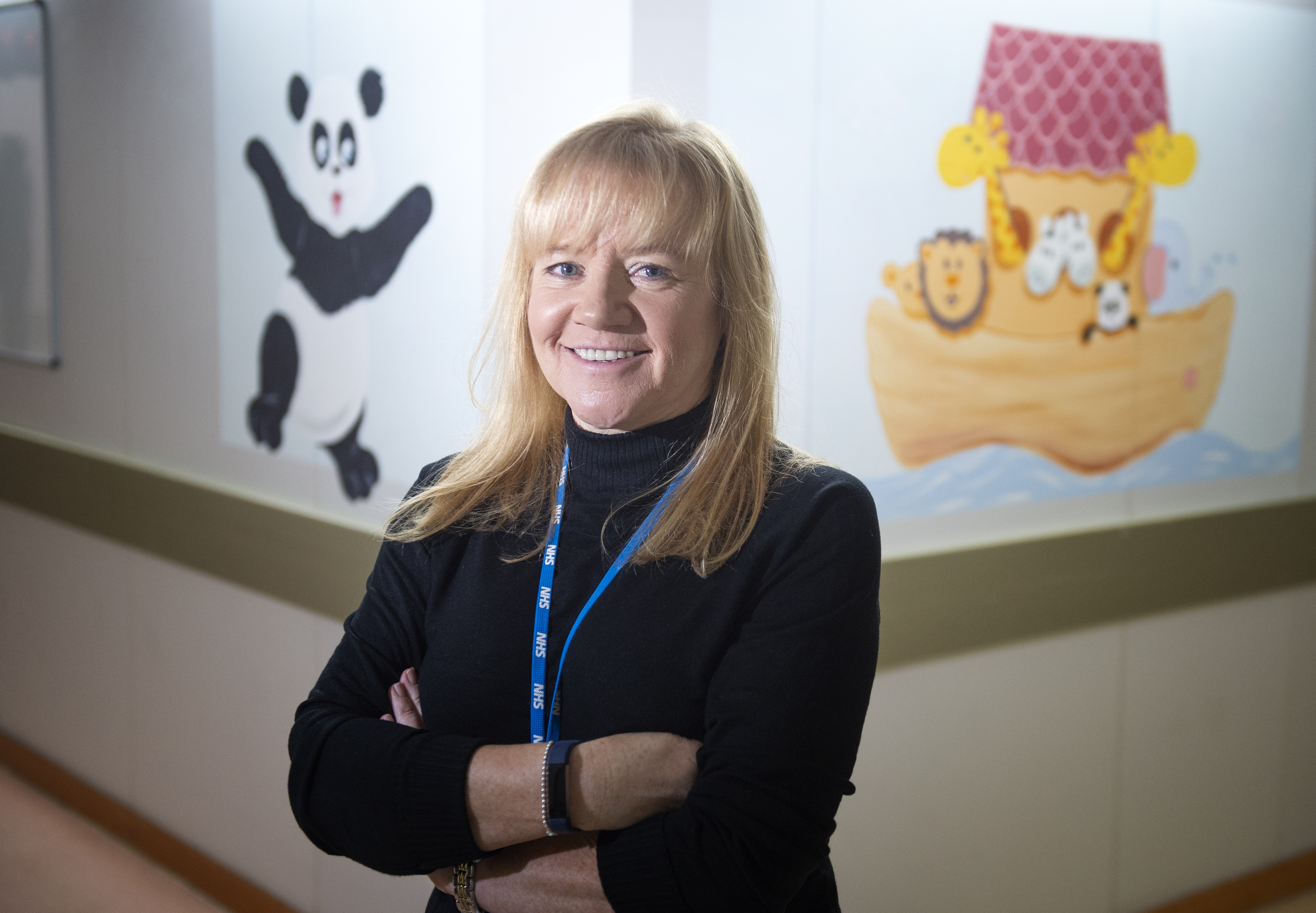 A person with long blonde hair and wearing a black long-sleeved top and a blue lanyard stands smiling with arms crossed. Behind them a wall with murals, one of a happy panda, the other a cartoon-style Noah’s ark with a panda, horses, giraffes and a lion visible.