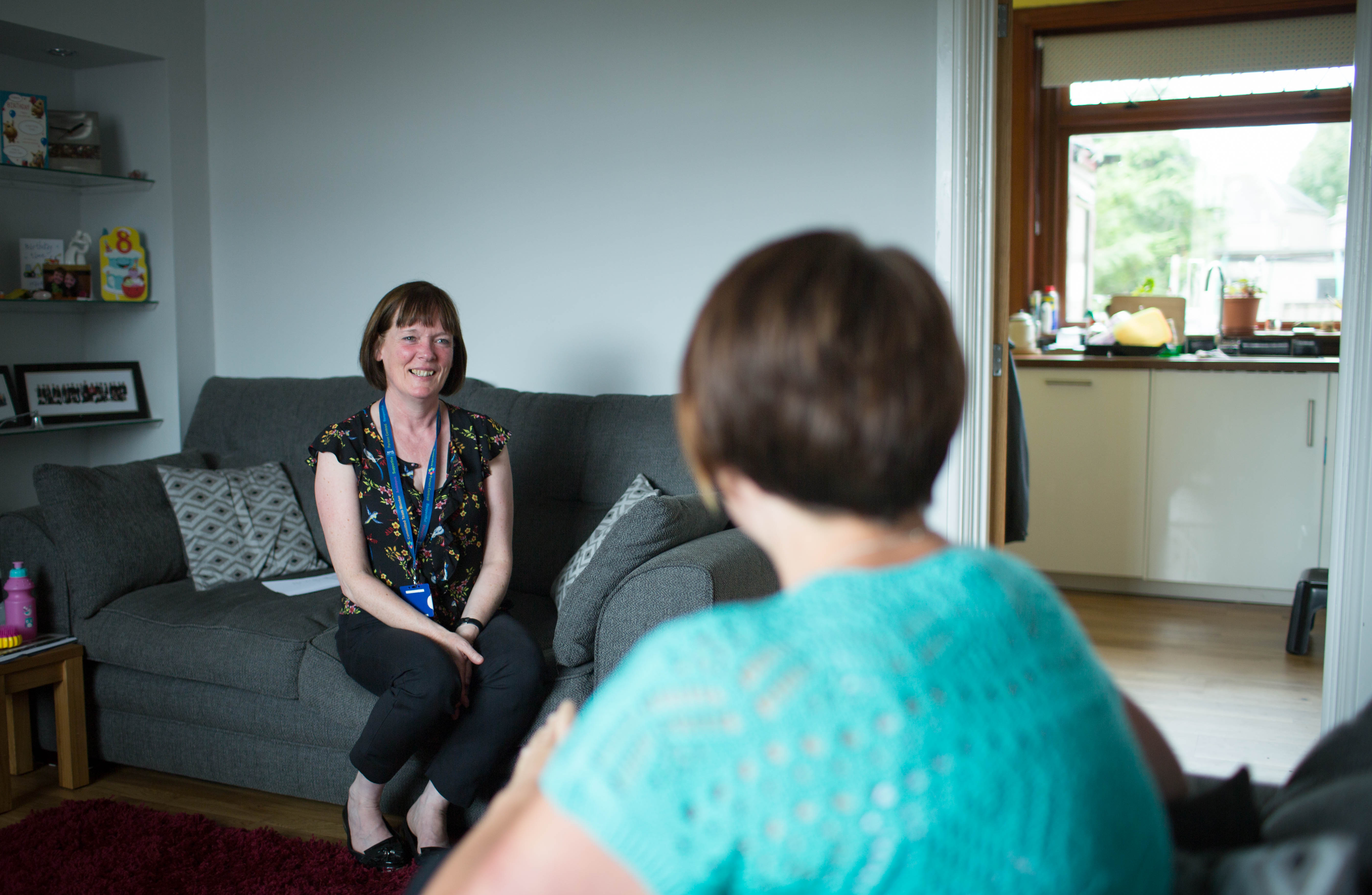 Two people sit in the lounge of a house facing each other. The person facing the camera is smiling. They have short dark hair and a fringe and are wearing a short sleeve black top with a pattern of flowers and birds. They're wearing a blue lanyard with a blue badge and black trousers and black shoes. They're sitting on a grey couch. The person closest to the camera has their back to it