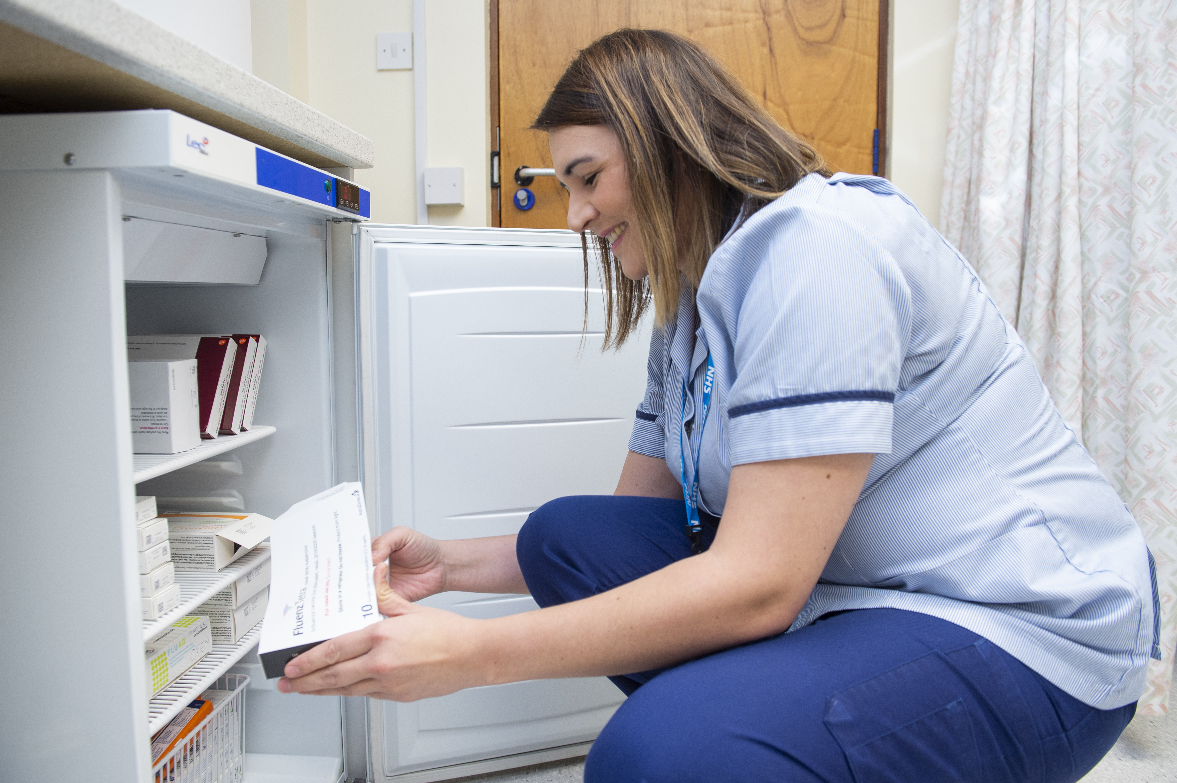 A healthcare worker in a light blue uniform is crouched in front of an open medical refrigerator, smiling while holding a box labeled "Fluarix Tetra." The fridge contains various medical supplies and vaccine boxes. The setting appears to be a clinical or healthcare environment.