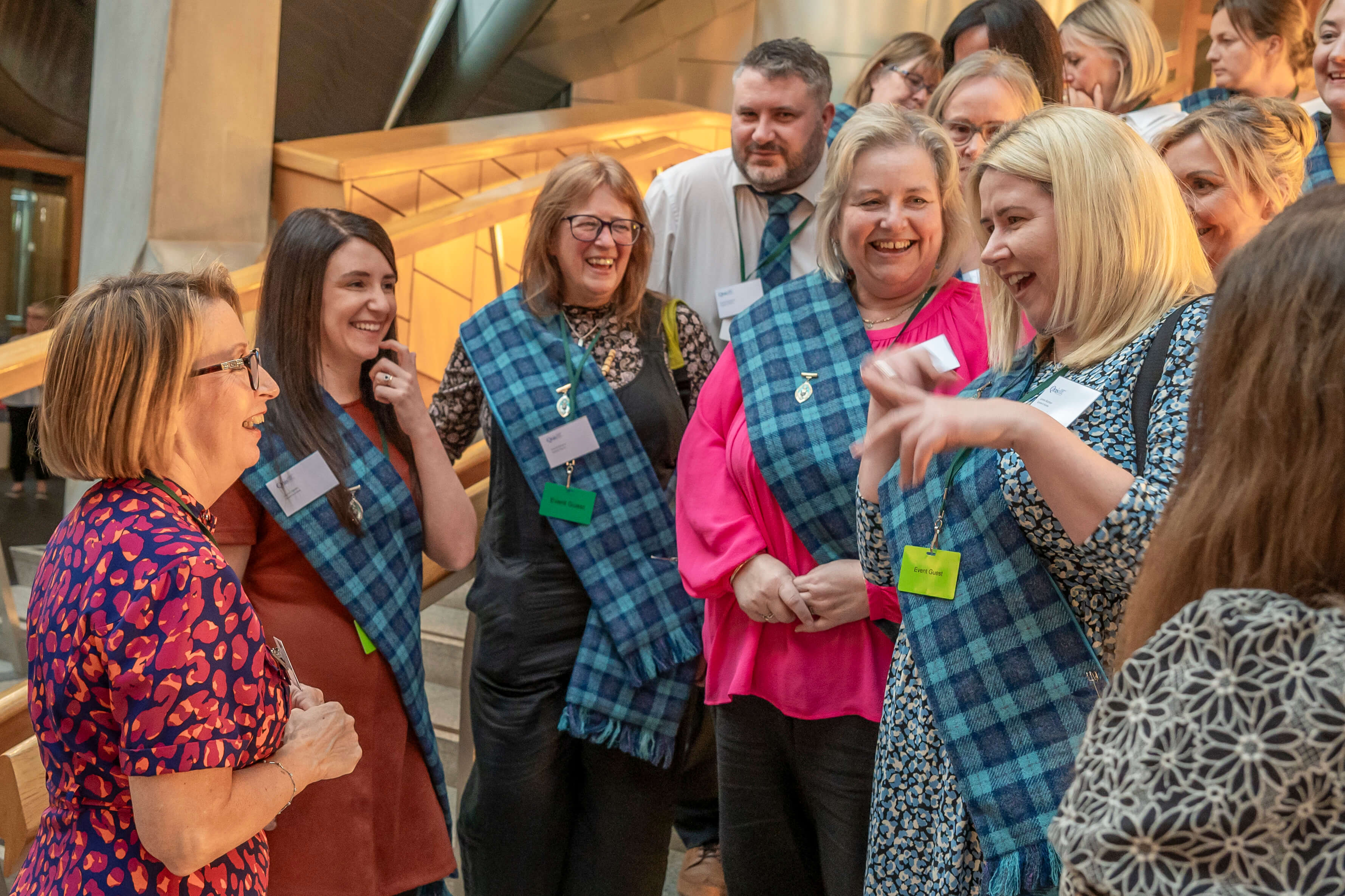 A group of people gathered indoors, smiling and talking animatedly. Several individuals are wearing blue tartan sashes over their clothing and have name badges and event passes around their necks. One person in the foreground wears a patterned top with red and purple shapes. The setting appears modern, with metal and wood architectural features in the background.
