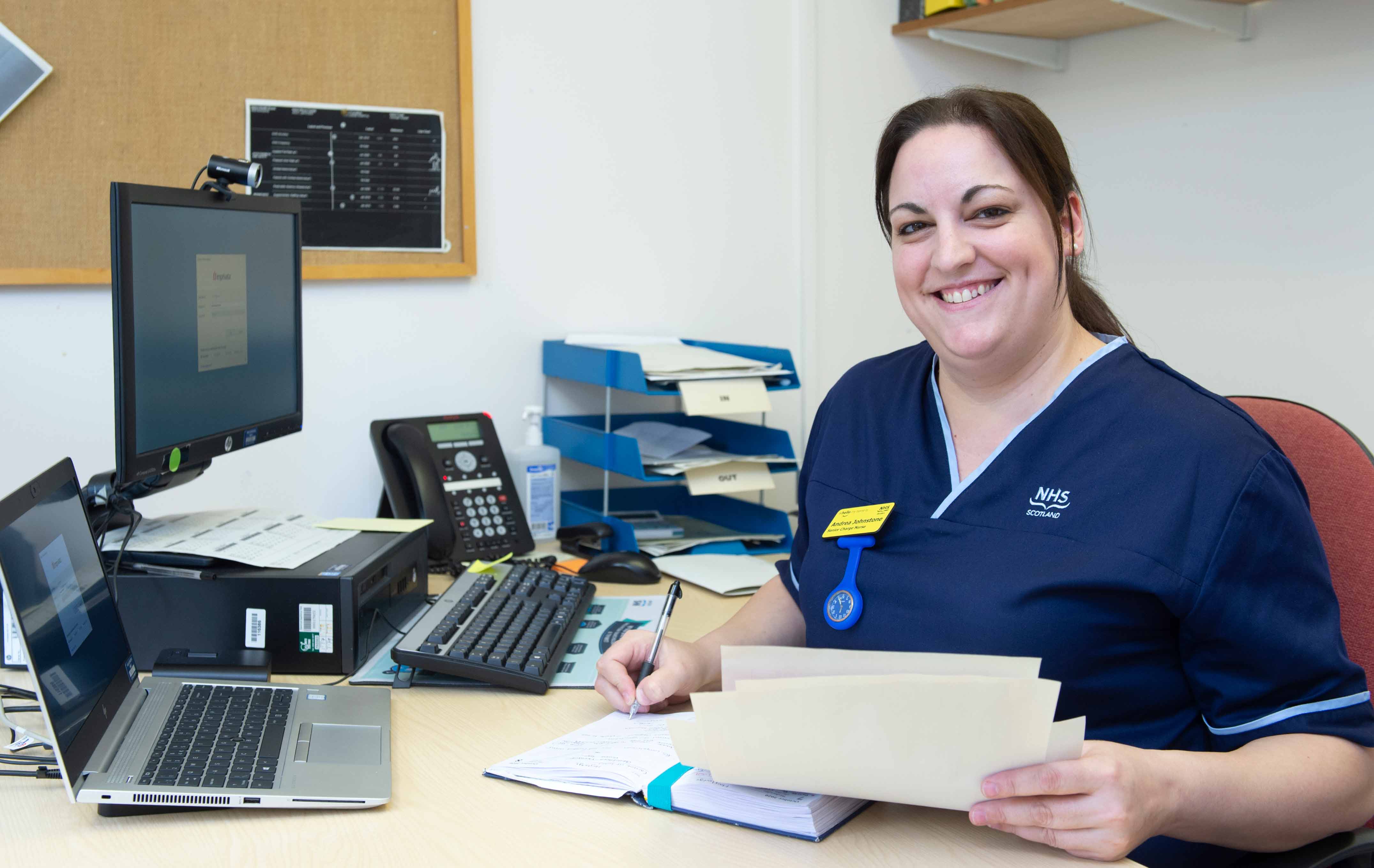 A dark-haired person wearing a navy blue NHS Scotland uniform sits at a desk, smiling at the camera. They hold a folder of papers in one hand and write in a notebook with the other. On the desk are a laptop, computer monitor, telephone, keyboard, and stacked trays for paperwork. A pinboard with pinned documents is visible on the wall behind.
