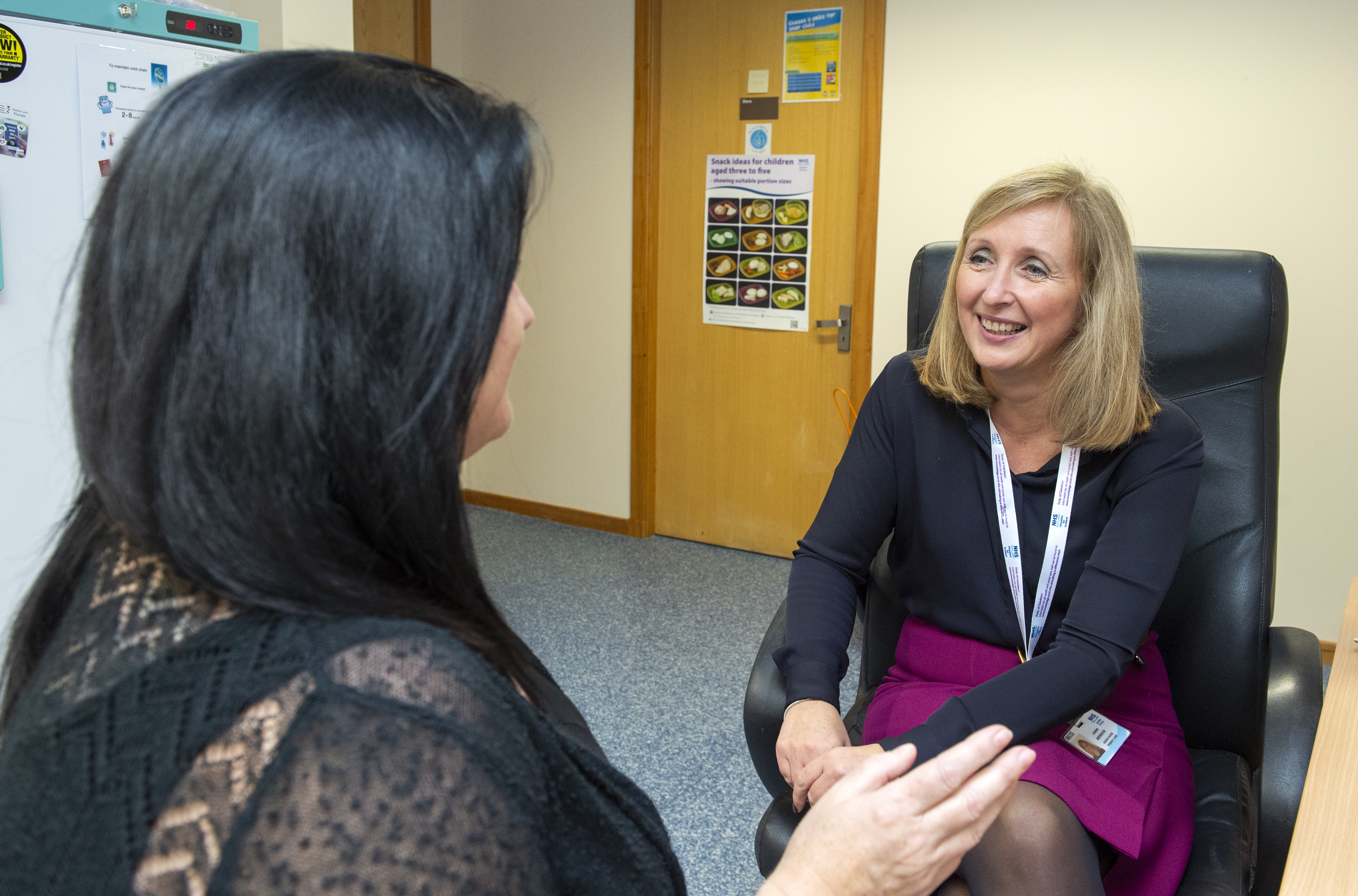 Two people sit across from each other. The person facing the camera smiles warmly, has shoulder length dark blonde hair, is wearing a black long sleeve top with a white lanyard, a knee length purple skirt and black tights. They're smiling at a person with their back to the camera, who has long black hair and is wearing a black lace top, they have a hand up as though talking animatedly. In the background, blue linoleum and a wooden door with signs stuck on it.