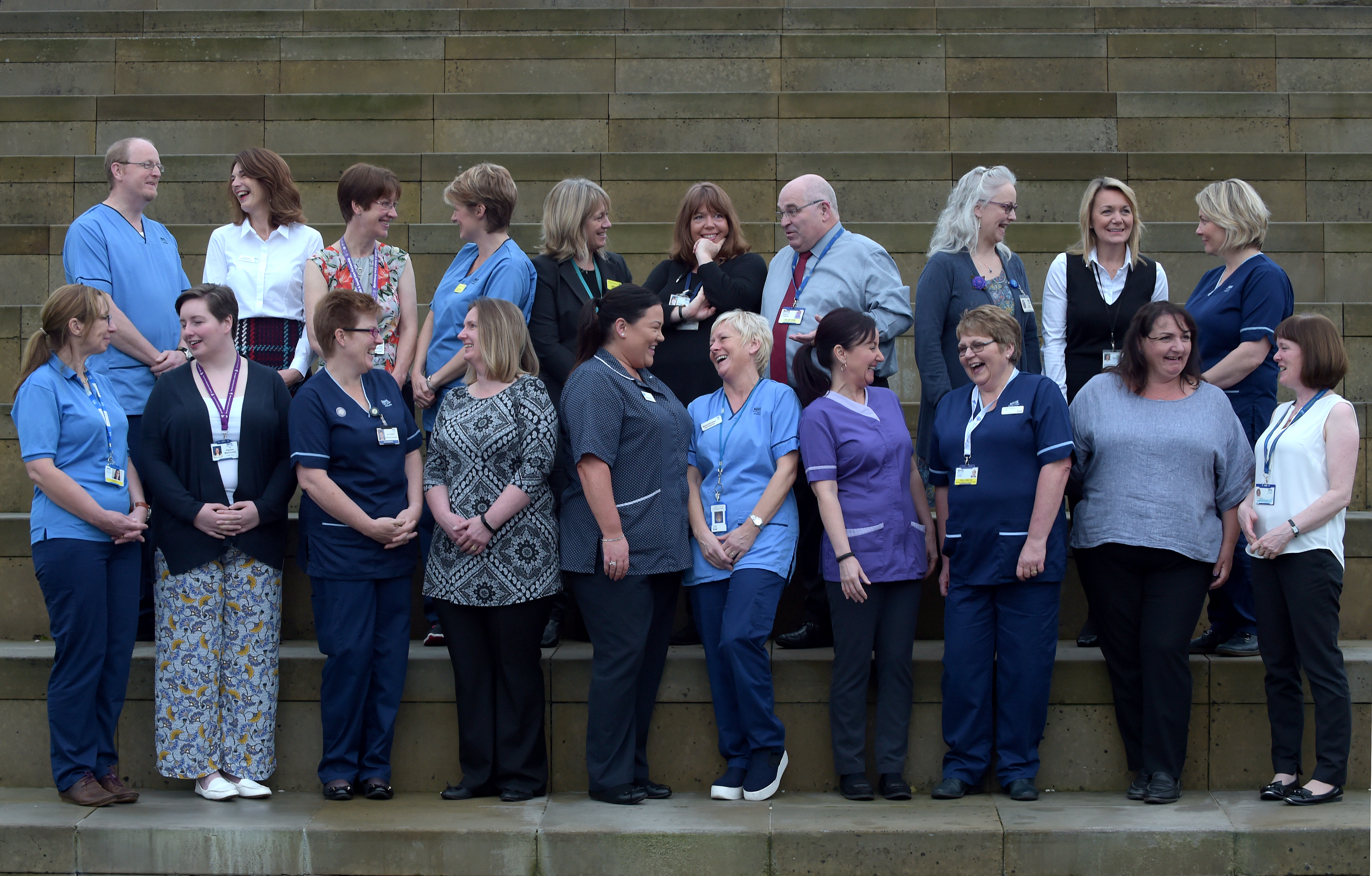 A group of people are gathered on a set of outdoor stone steps for a group photo. The group is composed of individuals wearing various uniforms, including blue medical scrubs, business casual attire, and ID badges. Most people are smiling or laughing, interacting with each other in a relaxed and friendly manner. The background features large, uniformly spaced stone steps.