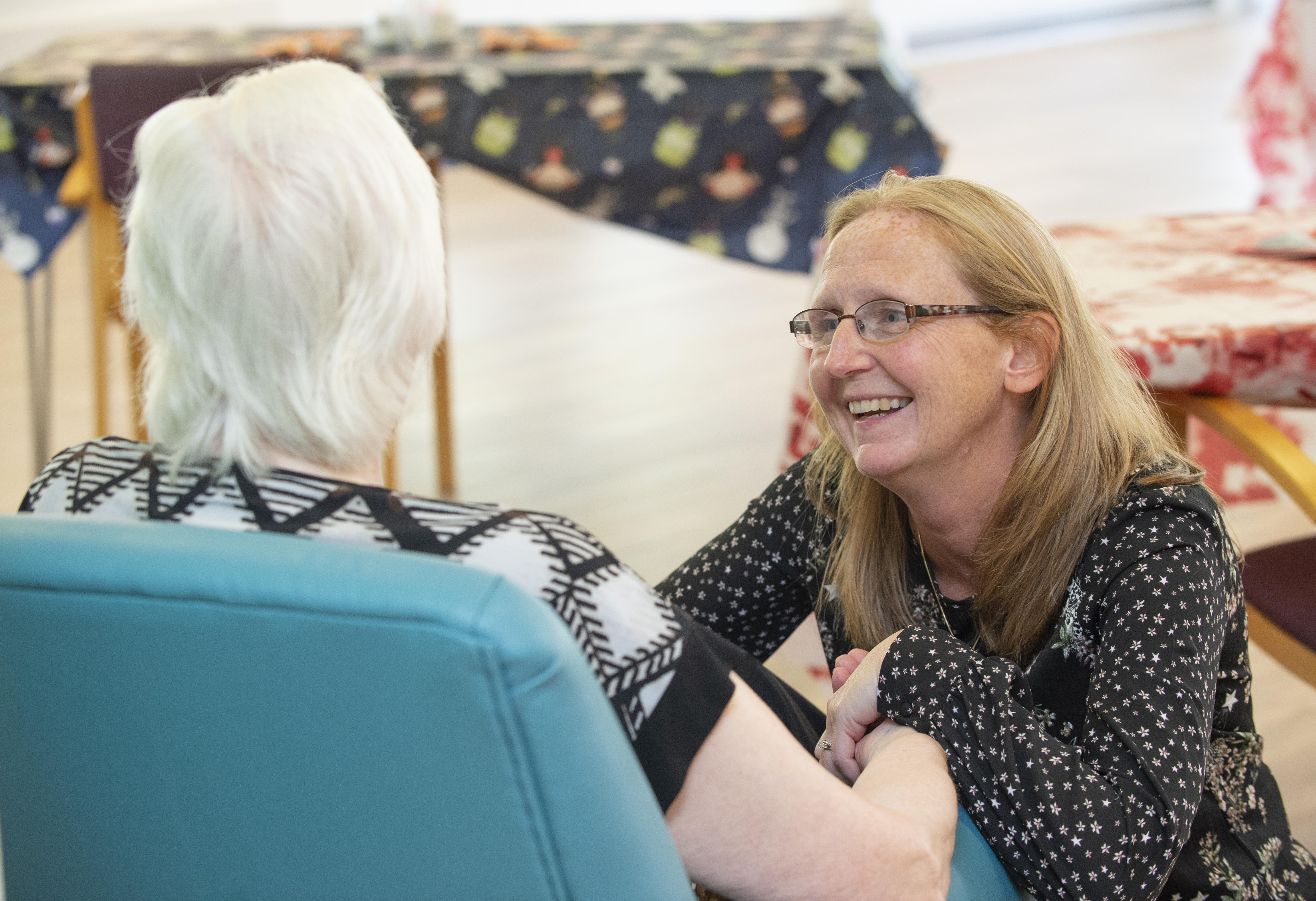 A smiling person with blonde hair below their shoulders wearing glasses and a black shirt with a white pattern of dots and flowers is crouched in front of a person sitting in in a turquoise chair. They have their back to the camera. They have short white hair and are wearing a geometric pattern white and black short sleeve top. In the background a table covered in a tablecloth and a red and white chair.