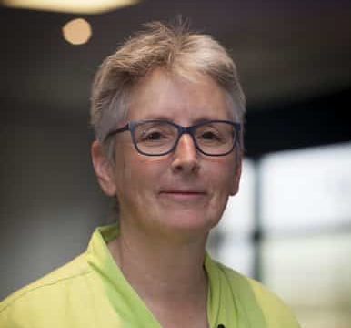 A person with short grey hair and glasses, wearing a light green collared shirt, standing indoors with soft lighting and a blurred background.