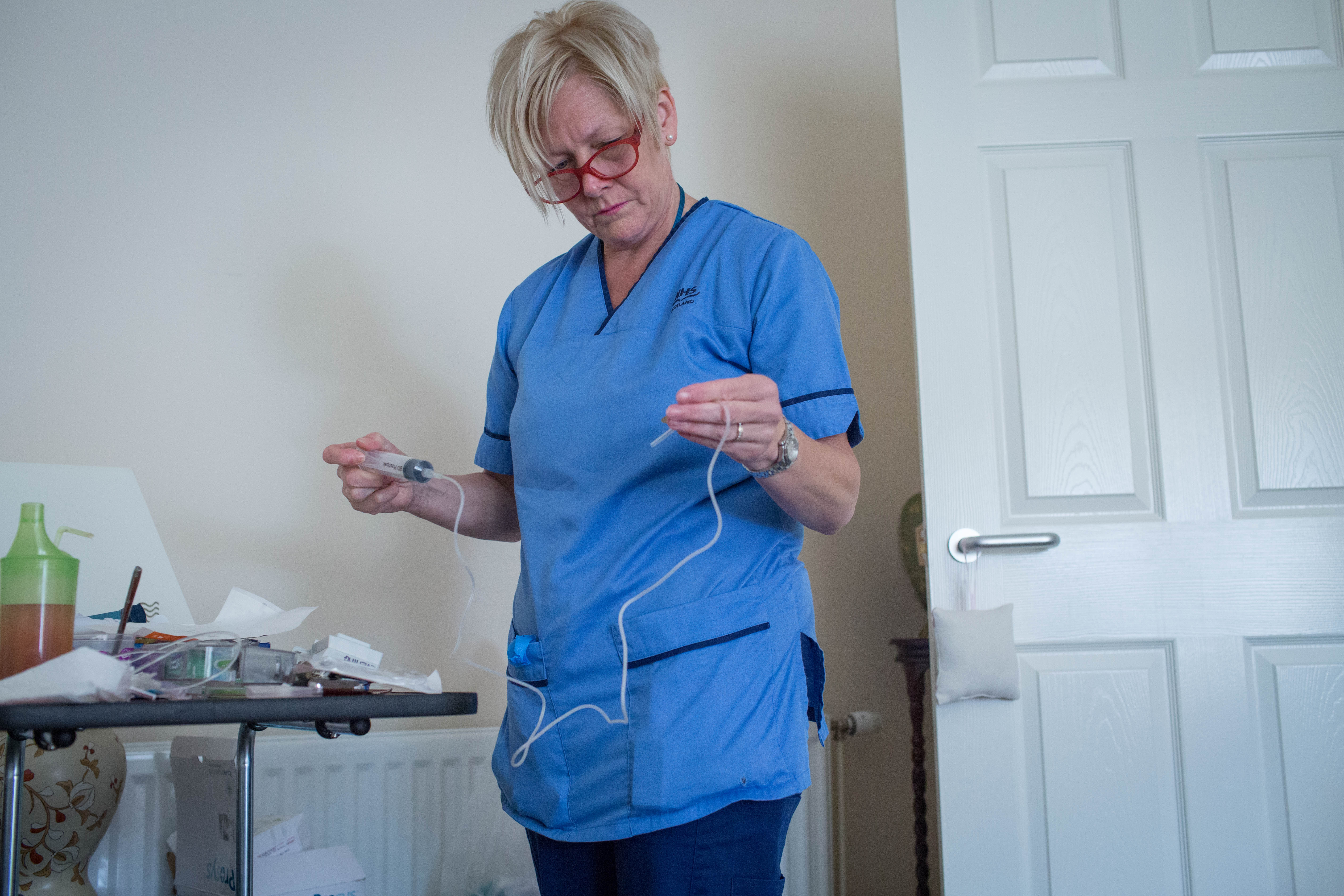 A person wearing a blue nurse’s uniform and red glasses is holding medical tubing and a syringe while standing in a room. They are focused on their task. A small table nearby holds various medical supplies, and a white door is open in the background.
