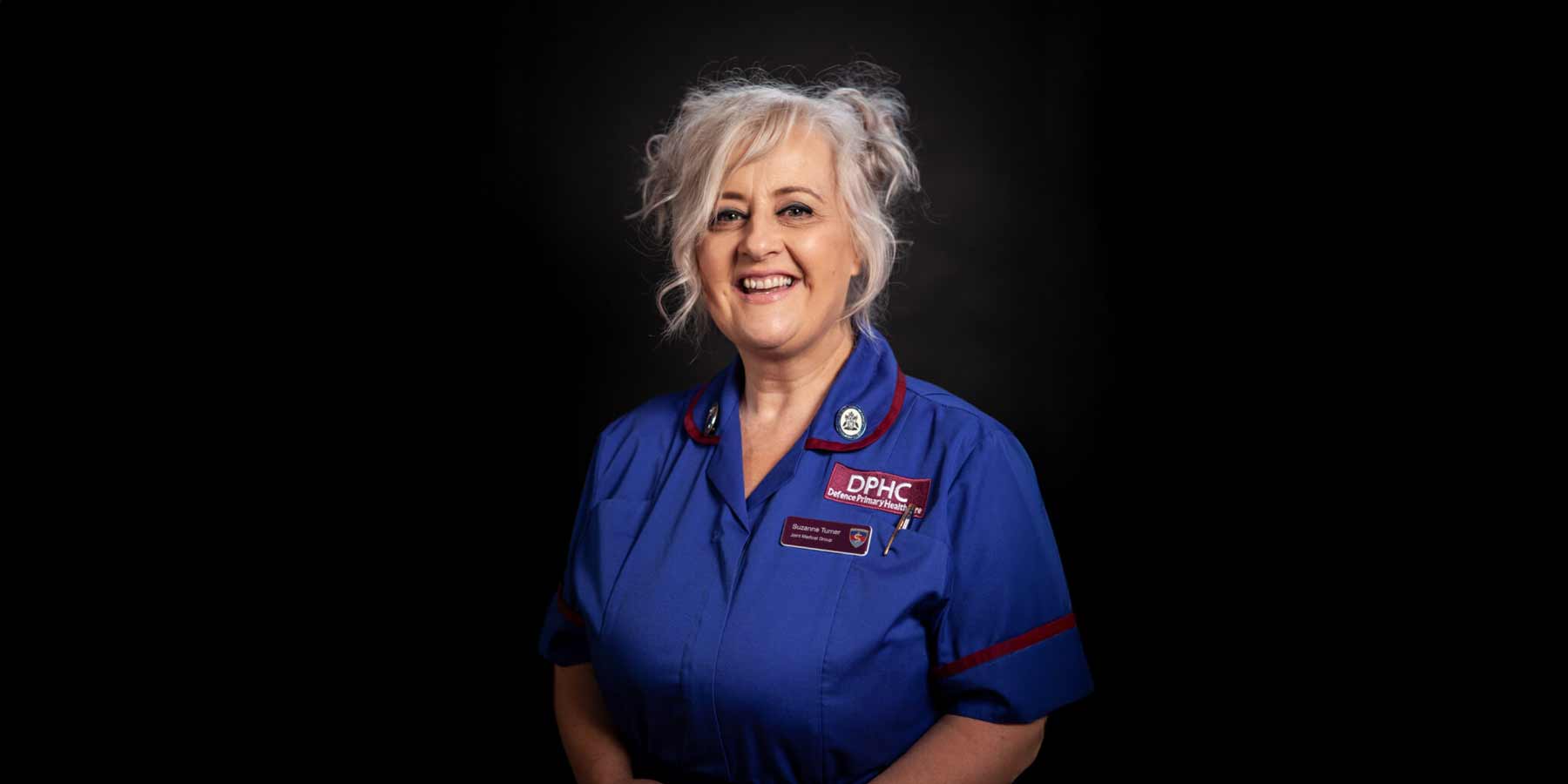 A person with light, curly hair styled in an updo smiles while wearing a dark blue medical uniform with maroon trim. The uniform has a badge reading 'DPhC Defence Primary Healthcare' and a name tag that says 'Suzanne Turner, Joint Medical Group.' The background is dark and plain.
