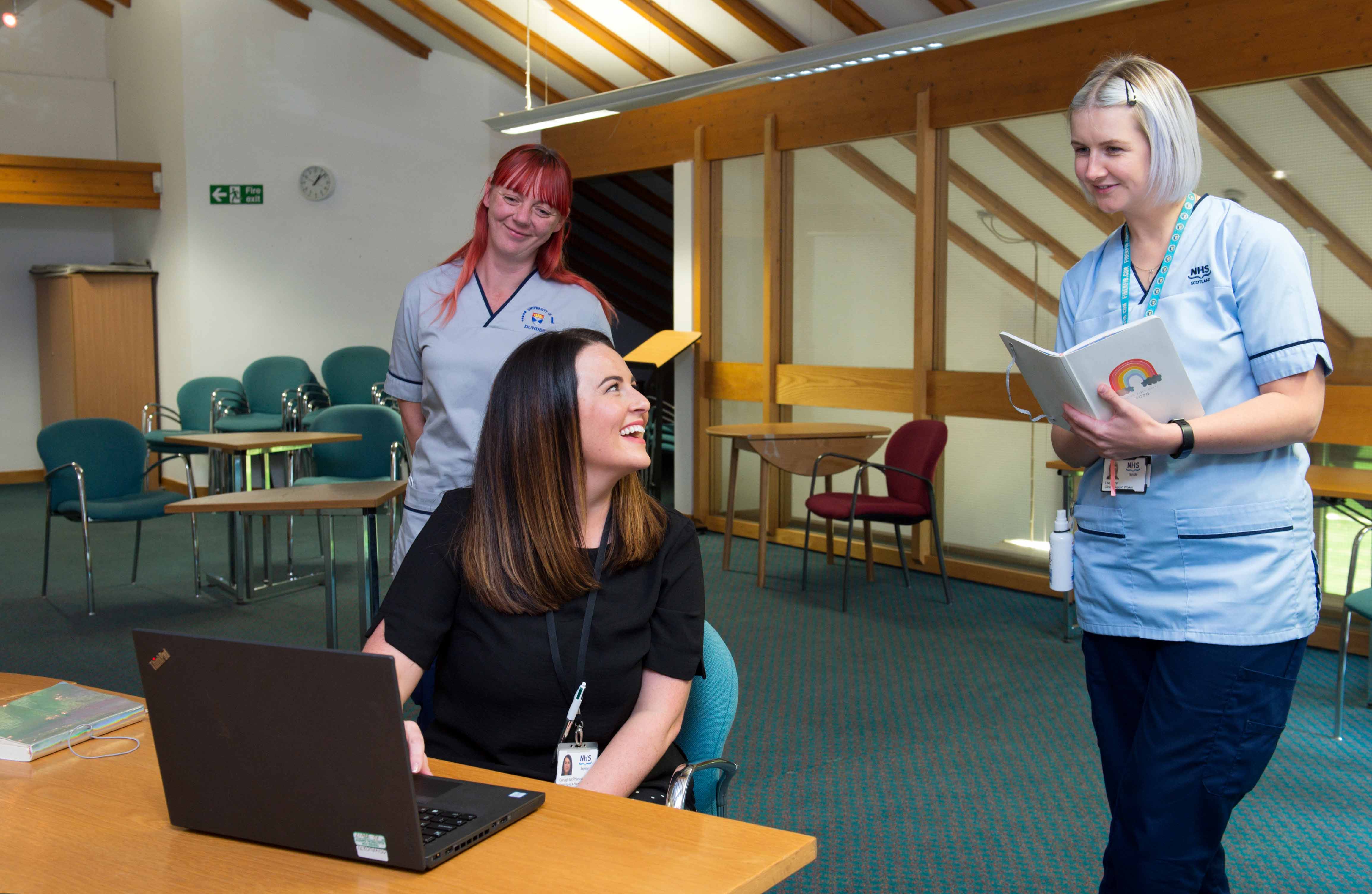 Three people in a brightly lit room with wooden beams. One person sits at a desk using a laptop and smiling at the two standing people, who are wearing NHS uniforms. One of the standing individuals is holding an open notebook with a rainbow sticker on the cover. There are empty chairs and tables in the background.