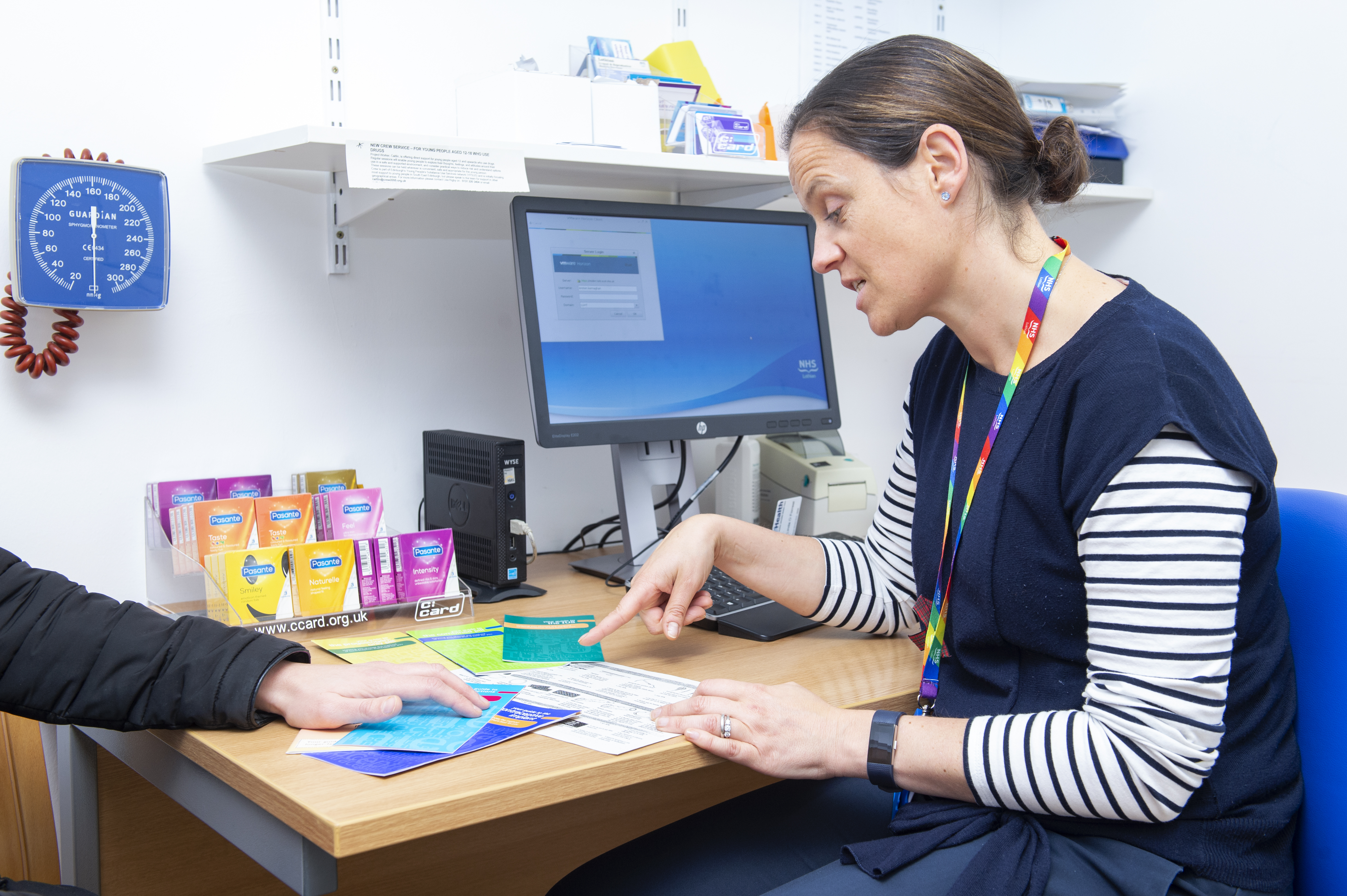 A healthcare professional is seated at a desk, engaged in conversation with another person whose arm is visible in the foreground. The professional is pointing to an informational leaflet on the desk, which is covered with various pamphlets and materials, including condom packs. A computer screen displaying an NHS login page is in the background, along with medical supplies and a blood pressure monitor on the wall. The setting appears to be a clinical consultation room.